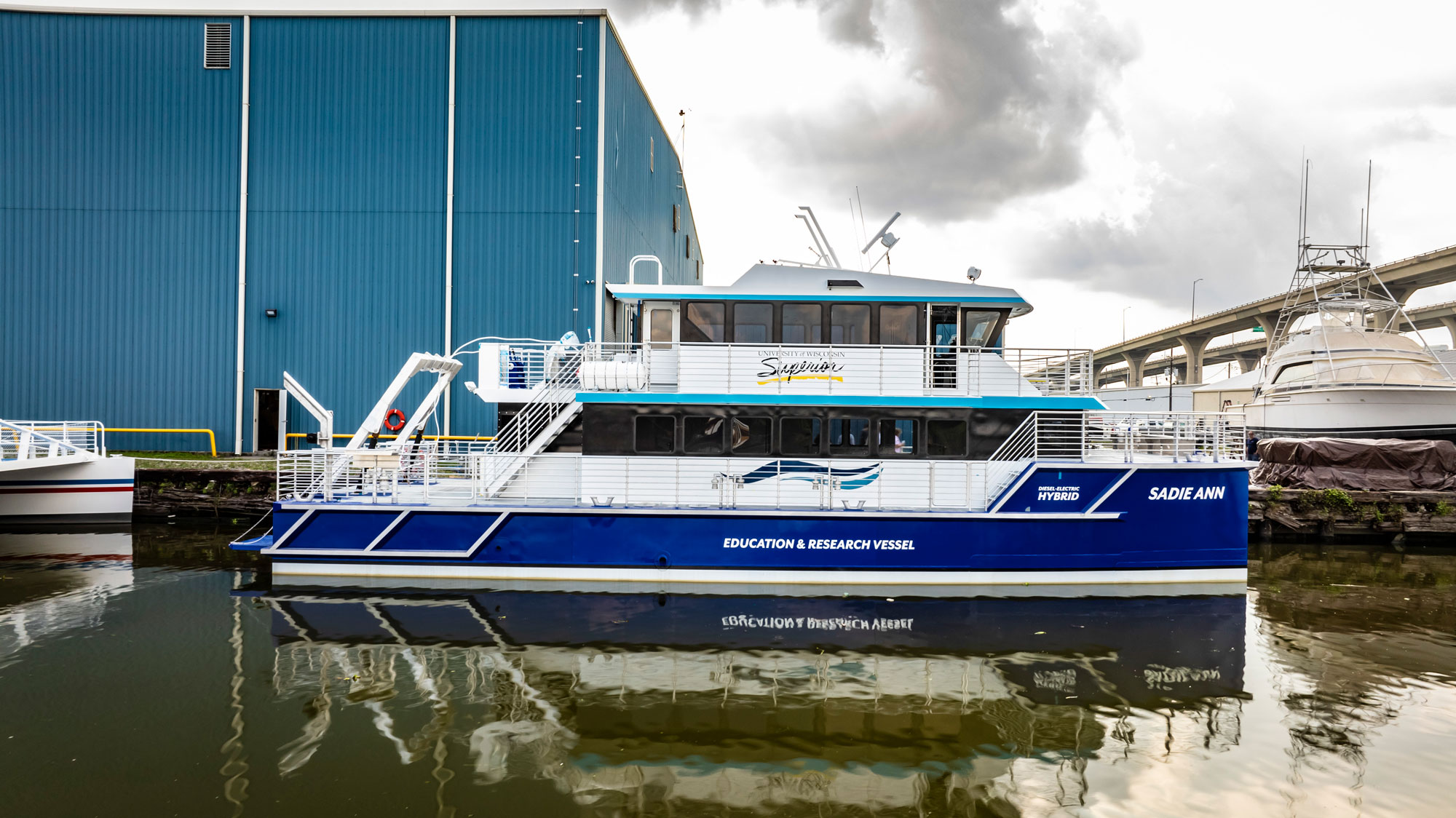 A blue and white education and research vessel named Sadie Ann is docked beside a blue warehouse, reflecting in the calm water under a cloudy sky—about to embark on its next voyage.