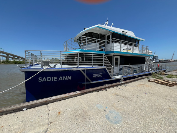 A blue and white boat named Sadie Ann is docked at a pier under a clear blue sky; the boat, with Hybrid and Silver signage on its side, is tied to the dock by a rope, inviting curiosity about its next journey.