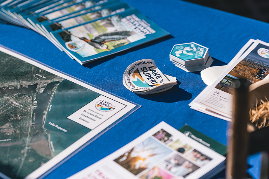 A table covered with a blue cloth displays maps, brochures, and various Lake Superior-themed stickers at an outdoor event.