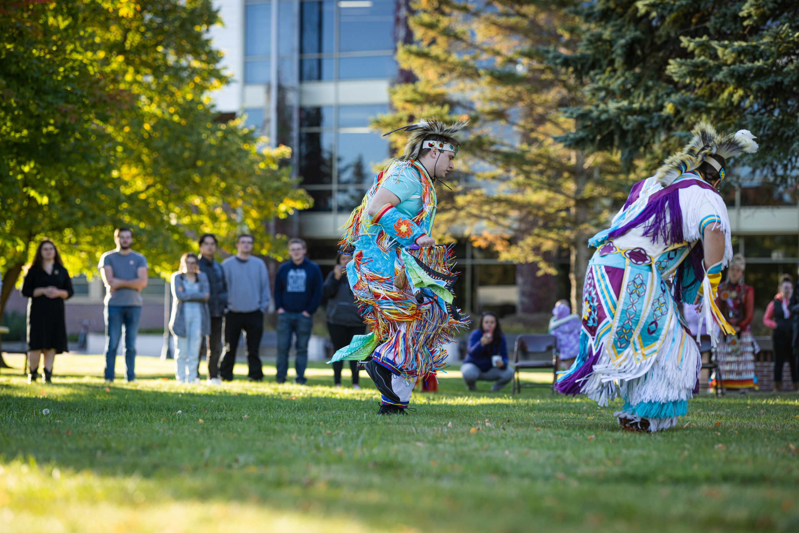 Two people in colorful traditional regalia perform a dance on grass, embodying equity, diversity, and inclusion. Onlookers watch near trees and a modern building as sunlight filters through, creating a vibrant outdoor scene.