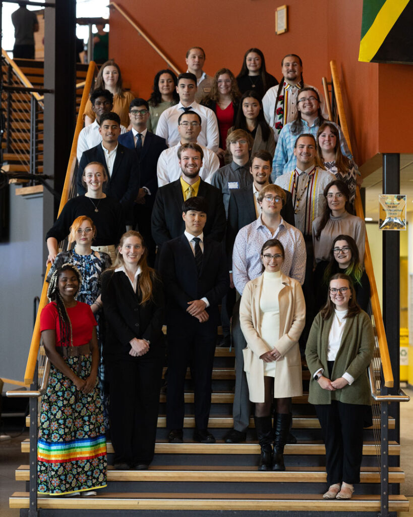 About 27 people stand in rows on a staircase indoors, all facing the camera. Most are dressed in business or semi-formal attire, while a few wear colorful traditional clothing, suggesting the setting is a professional or academic event.