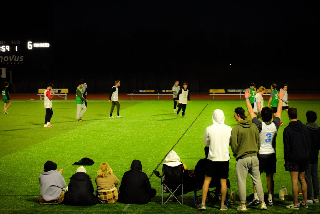 A group of people, some seated and some standing, watch an evening soccer game on a brightly lit field. Players in green and white jerseys are on the field, and a scoreboard is visible in the background.
