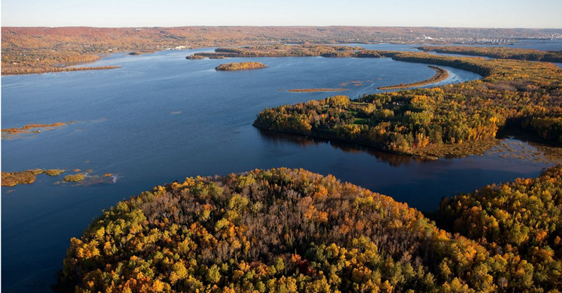 Aerial view of a winding river surrounded by islands and forests with autumn foliage in shades of green, yellow, and orange under a clear sky, capturing the beauty about nature’s seasonal transformation.
