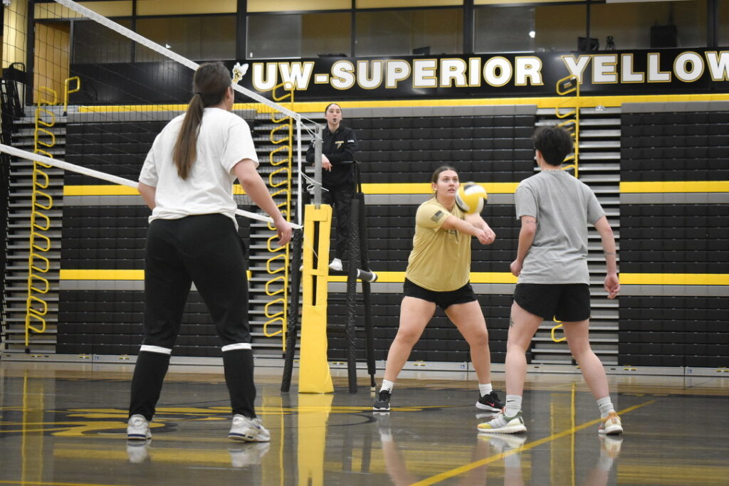 Three volleyball players are on an indoor court; one is hitting the ball with a forearm pass while the others watch. An official stands on a referee stand in the background. The gym has yellow and black bleachers.
