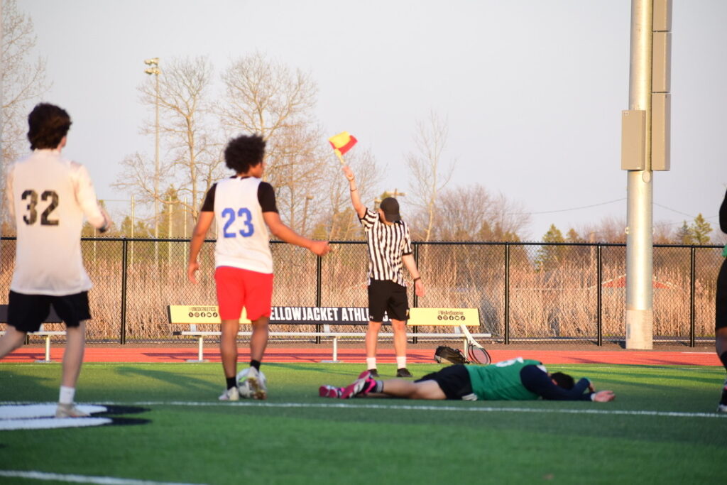 A referee holds up a yellow and red flag on a sports field while a player in green lies on the ground and other players in jerseys with numbers walk nearby. Trees and a bench are visible in the background.