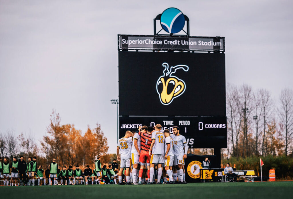Athletics-(3) A soccer team in yellow and white uniforms huddles on the field in front of a large scoreboard at SuperiorChoice Credit Union Stadium, with players and trees visible in the background.
