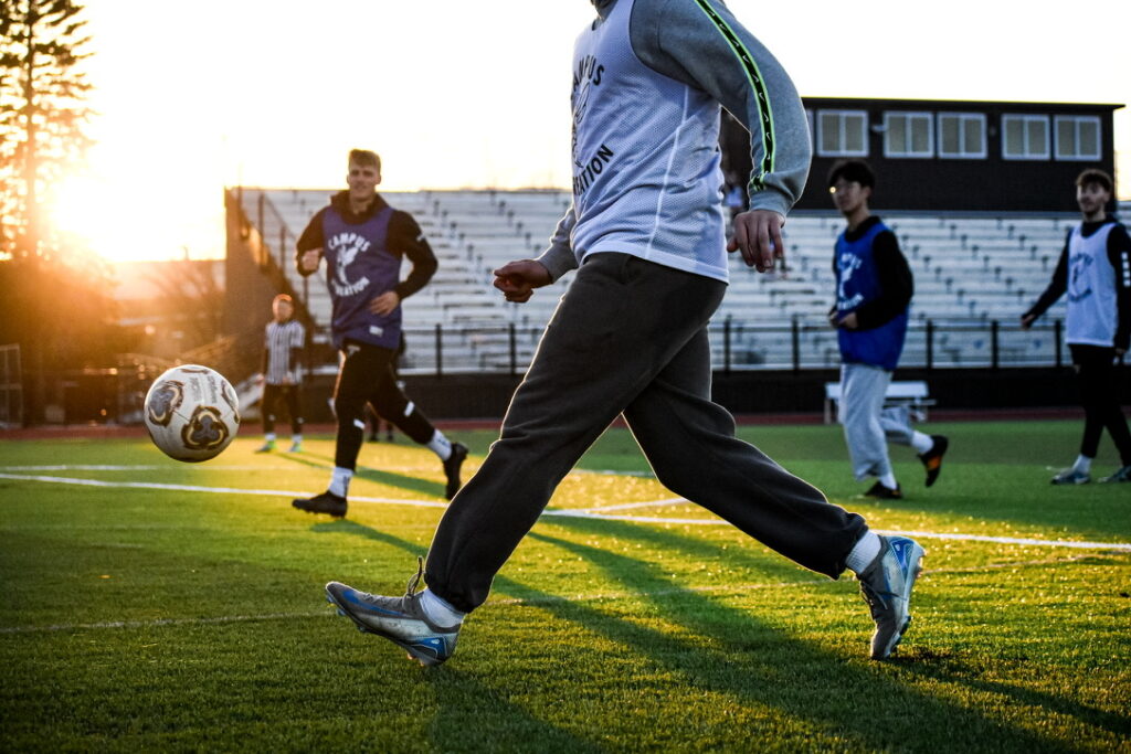 A group of people play soccer on a field at sunset. One person in the foreground is about to kick the ball, while others watch and run in the background near empty bleachers.
