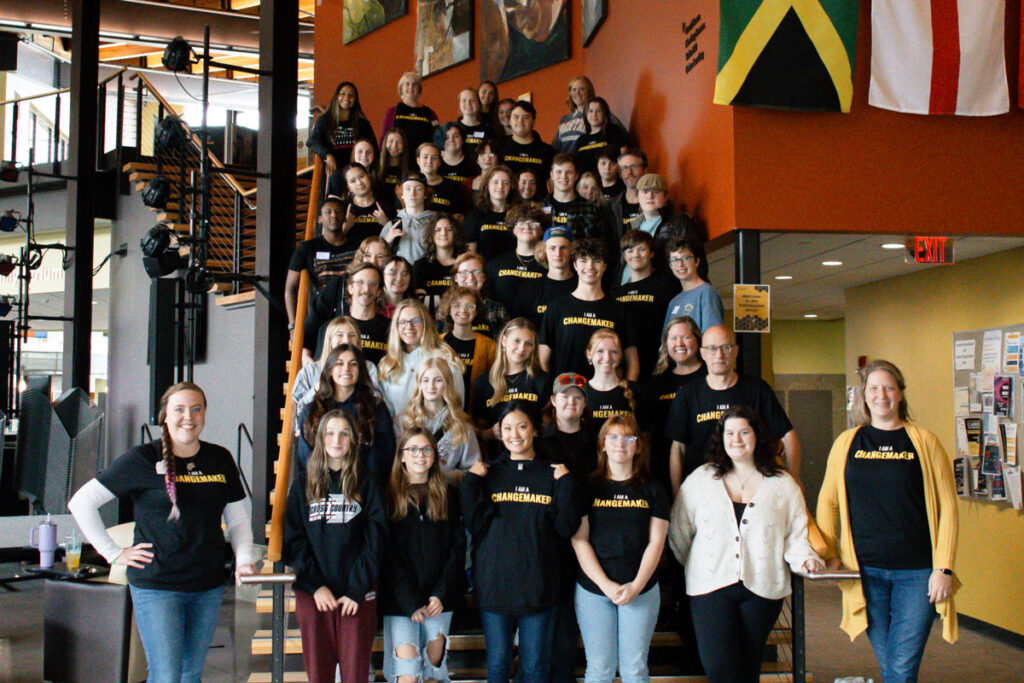 A large group of people, mostly teens and a few adults, pose on a staircase inside a building during Summer Camp. Many wear matching black Changemaker shirts. There are flags and art on the walls and posters on a bulletin board.