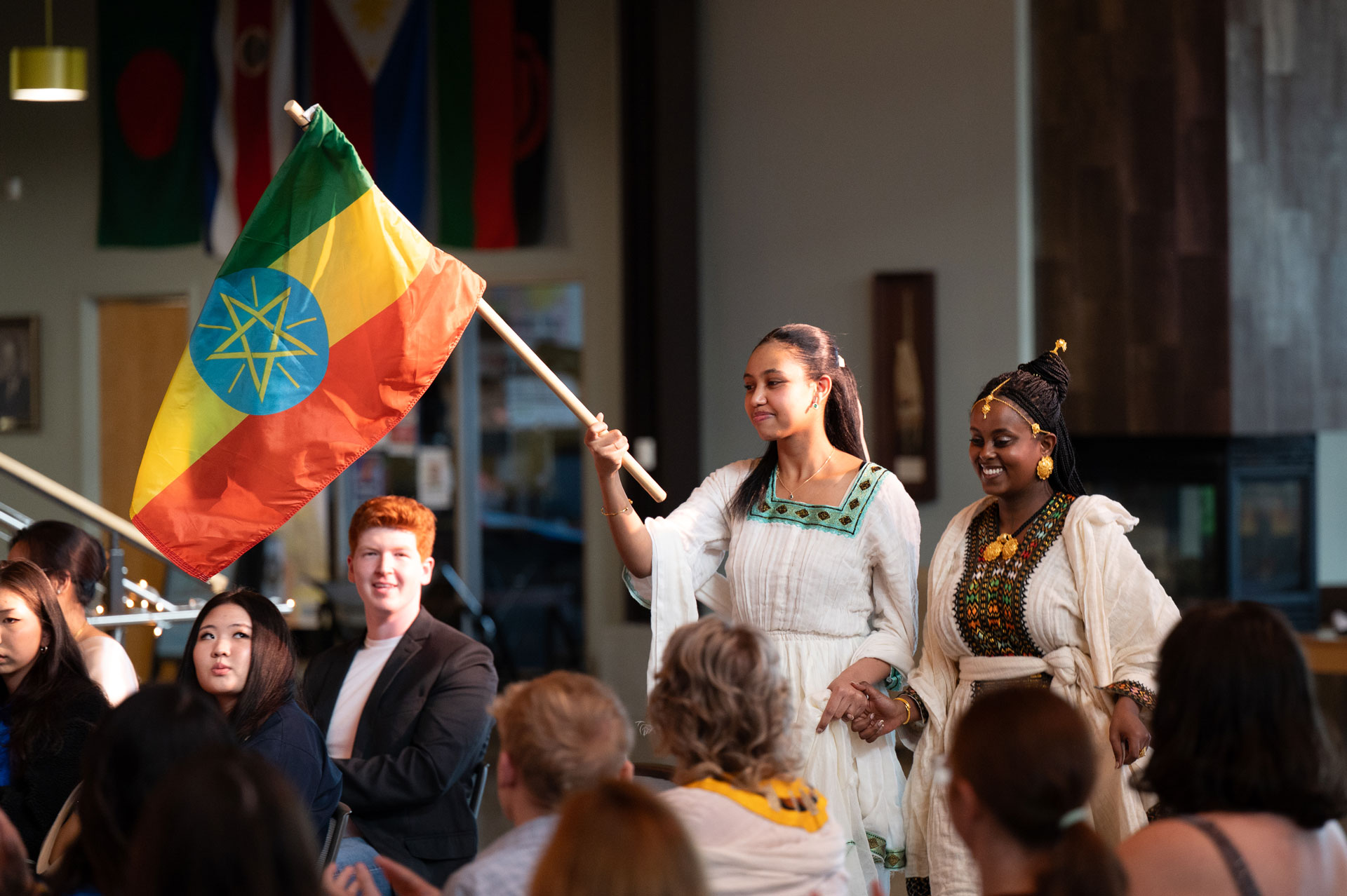 Two women in traditional Ethiopian clothing walk through a crowd, one holding the Ethiopian flag. People watch and smile, with various national flags in the background, inspiring others to get involved in the bright, celebratory atmosphere.