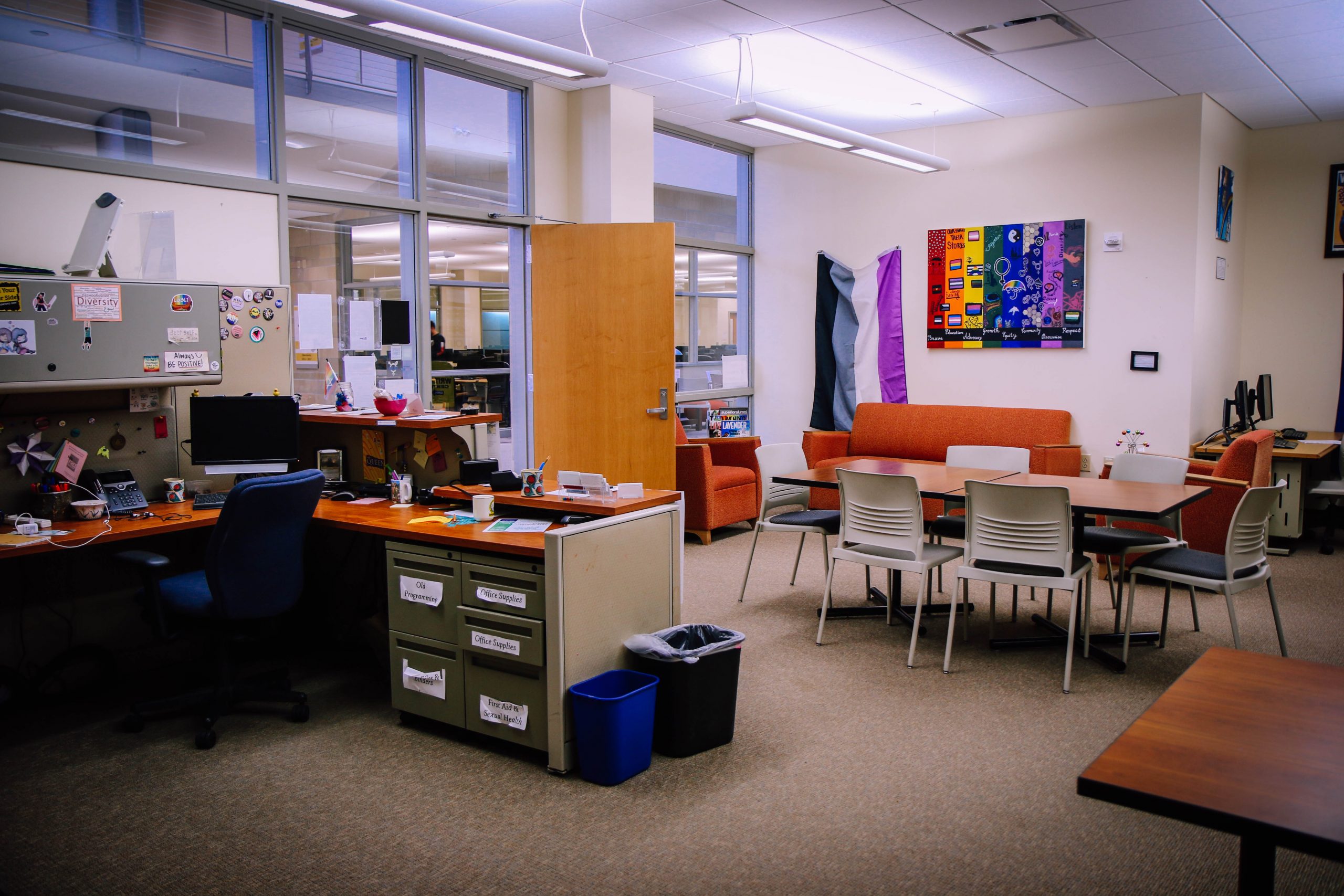 A modern office space with desks, computers, filing cabinets, and a meeting area with chairs and a table. Orange couches and colorful artwork decorate the room. Large windows let in natural light.
