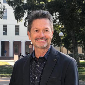 A man with short, styled hair and a goatee, wearing a dark blazer and patterned shirt, smiles while standing outdoors in front of a building with large windows—an area often lively with past events and activities.