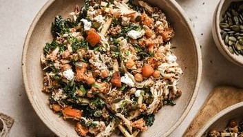 A bowl filled with a colorful salad containing kale, roasted sweet potatoes, chickpeas, quinoa, seeds, and crumbled cheese, placed on a light-colored surface.