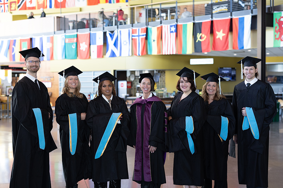 A group of six graduates in caps and gowns and one faculty member in academic regalia stand smiling in a campus building, with international flags hanging in the background.