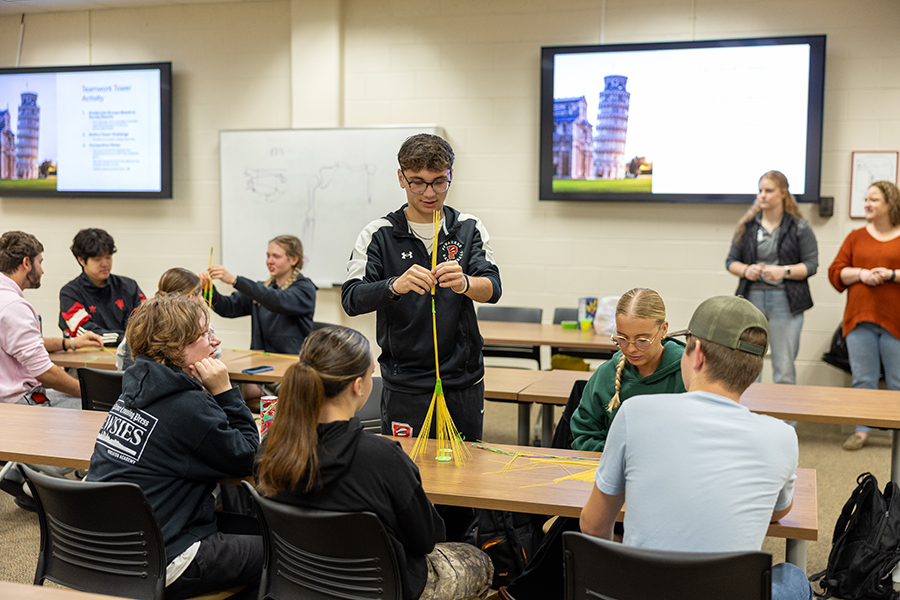 Students sit in groups at tables in a classroom, working together to build structures with uncooked spaghetti and tape. A presentation with an image of the Leaning Tower of Pisa is displayed on a screen in the background.