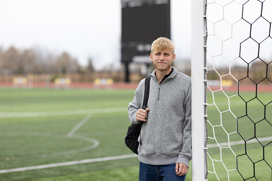 A young man with blonde hair stands by a soccer goalpost on a field, wearing a gray pullover and carrying a black bag over his shoulder. The background shows a blurred scoreboard and trees.