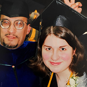 A smiling young woman in a graduation cap and gown poses with an older man in academic regalia, who playfully makes bunny ears behind her head.