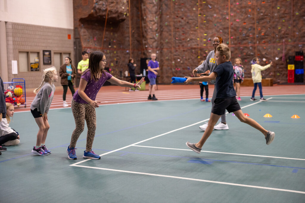 Children participate in a relay race inside a gym at summer camp, with one child handing a baton to another. Other kids and adults watch, and a climbing wall is visible in the background.
