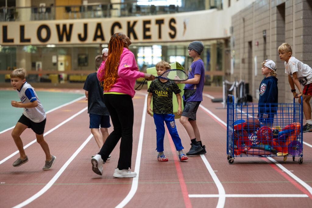 A group of children at a Youth Camp stand on an indoor track near a woman with long red hair, while some kids run and others gather by a cart full of sports equipment. The word YELLOWJACKETS is visible in the background.