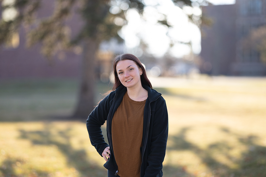 A young woman with long brown hair, wearing a brown shirt and black jacket, stands outdoors on a sunny day with trees and a building blurred in the background. She is smiling and has one hand on her hip.