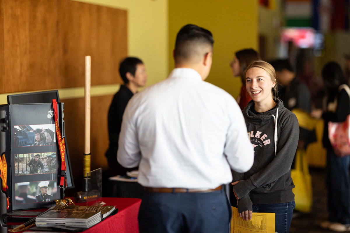 A young woman smiles while talking to a man at a career fair. Photos and event information are displayed on a nearby table, while other people interact in the background.