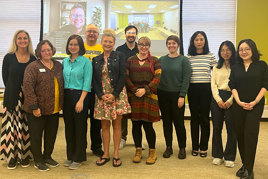 A group of twelve people stand indoors, posing and smiling for a group photo. Behind them, a screen shows a virtual meeting with four participants visible. The room has bright lighting and neutral-colored walls.