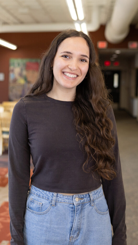 A young woman with long, wavy brown hair smiles while standing indoors. She is wearing a black long-sleeve shirt and light blue jeans. The background includes tables, chairs, and a hallway.
