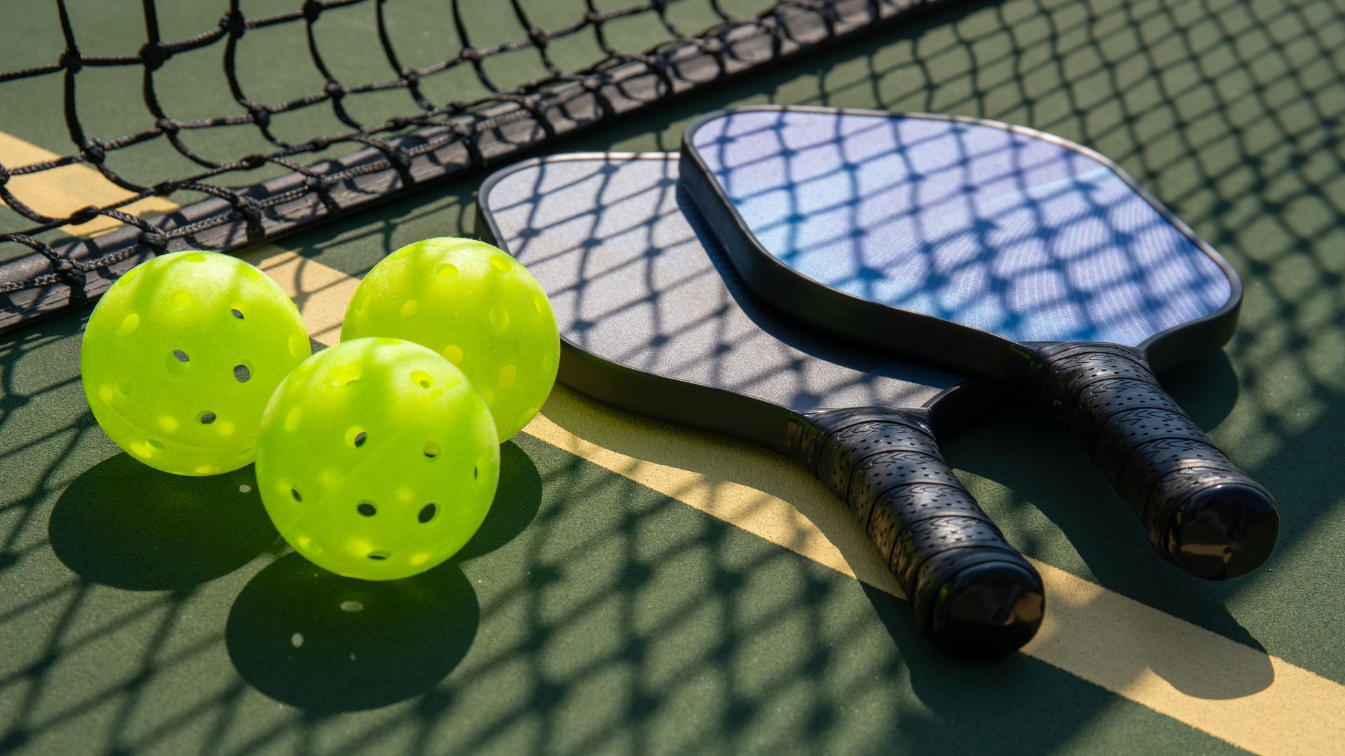 Two pickleball paddles and three bright yellow pickleballs rest on an outdoor court near the net, with shadows from the net visible on the equipment and surface.