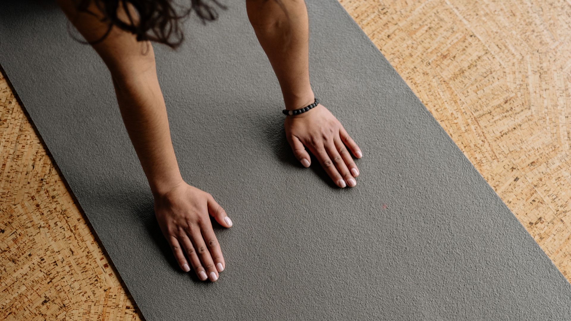 A person’s hands and forearms are pressed on a gray yoga mat, positioned as if starting a push-up, with a wooden textured floor in the background.
