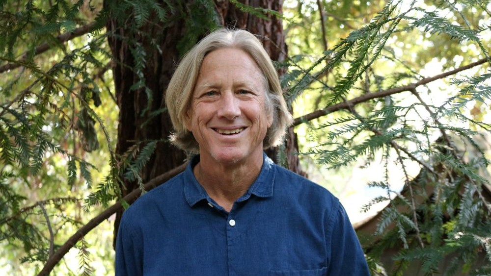 A smiling person with shoulder-length blonde hair stands outdoors in front of a tree and green foliage, wearing a blue button-up shirt.