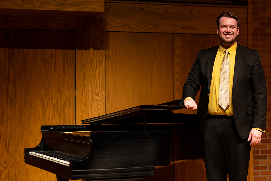 A man in a black suit with a yellow shirt and patterned tie stands smiling beside a grand piano in a wood-paneled room.