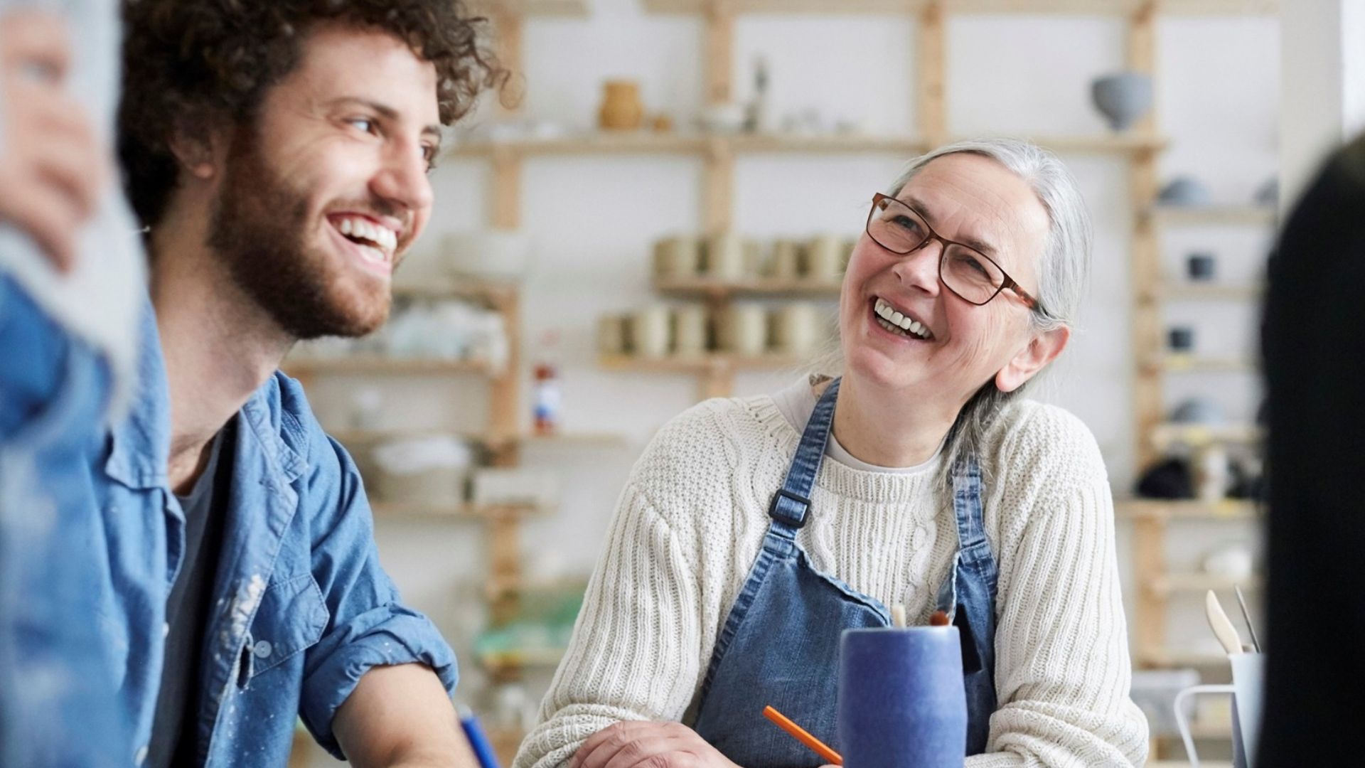 Two people, a younger man with curly hair and a woman with gray hair and glasses, smile and laugh while sitting at a table in a brightly lit studio with shelves of pottery in the background.