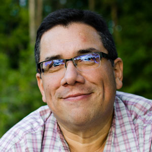 A middle-aged man with short dark hair and glasses is smiling, wearing a plaid shirt. The background of blurred greenery hints at past events and activities enjoyed outdoors.