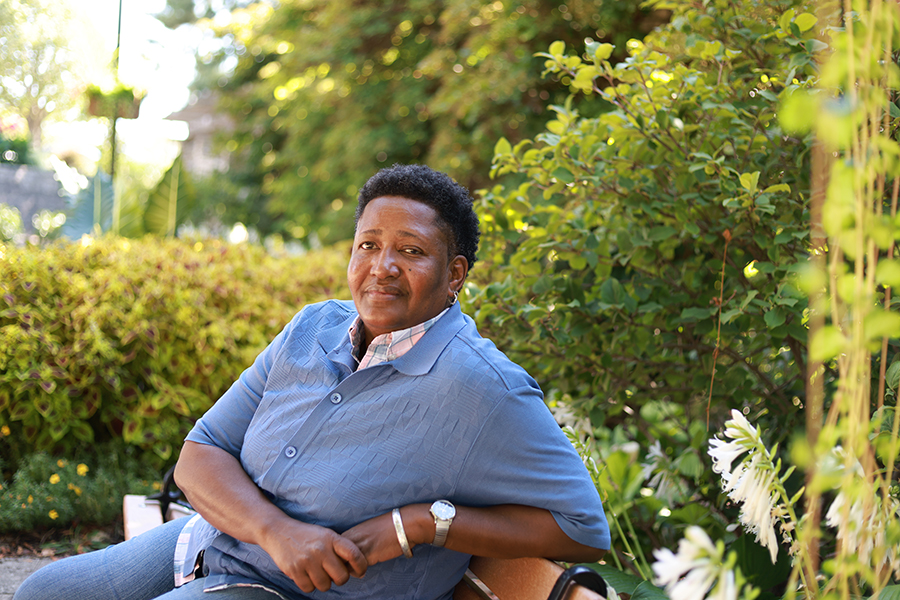 A person wearing a blue shirt sits on a bench in a garden, surrounded by green foliage and white flowers, looking calmly at the camera. Sunlight filters through the trees in the background.