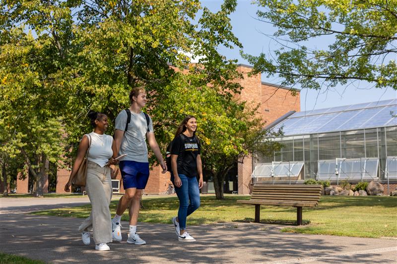 Three students walk together on a sunny campus path, chatting and smiling, with trees, a bench, and a brick building with a glass greenhouse in the background.