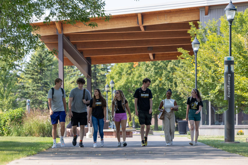 A group of seven University of Wisconsin-Superior students walk together on a tree-lined campus path under a wooden canopy, talking and smiling on a sunny day. A blue emergency call box is visible nearby.