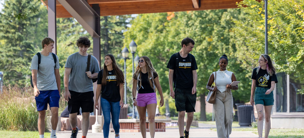 A group of six University of Wisconsin-Superior students walk together outdoors on a sunny day, wearing casual clothes and backpacks, with trees and campus buildings in the background.