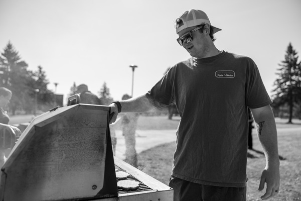 A man wearing sunglasses and a backwards cap grills food outdoors in a park, with smoke rising from the barbecue and trees visible in the background, enjoying a cookout after moving to campus.