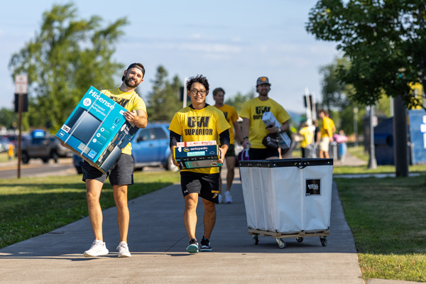 A group of people in yellow shirts walk on a sidewalk carrying boxes and pushing a large laundry cart, likely helping with moving or unloading during the Weekend of Welcome. Trees and parked cars are visible in the background.