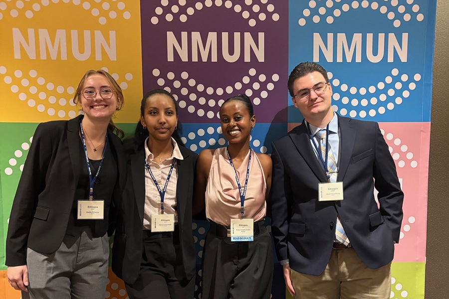 Four people dressed in business attire stand and smile in front of a colorful backdrop with the letters “NMUN” repeated in white circles on yellow, green, purple, and blue backgrounds. They are wearing name tags and lanyards.