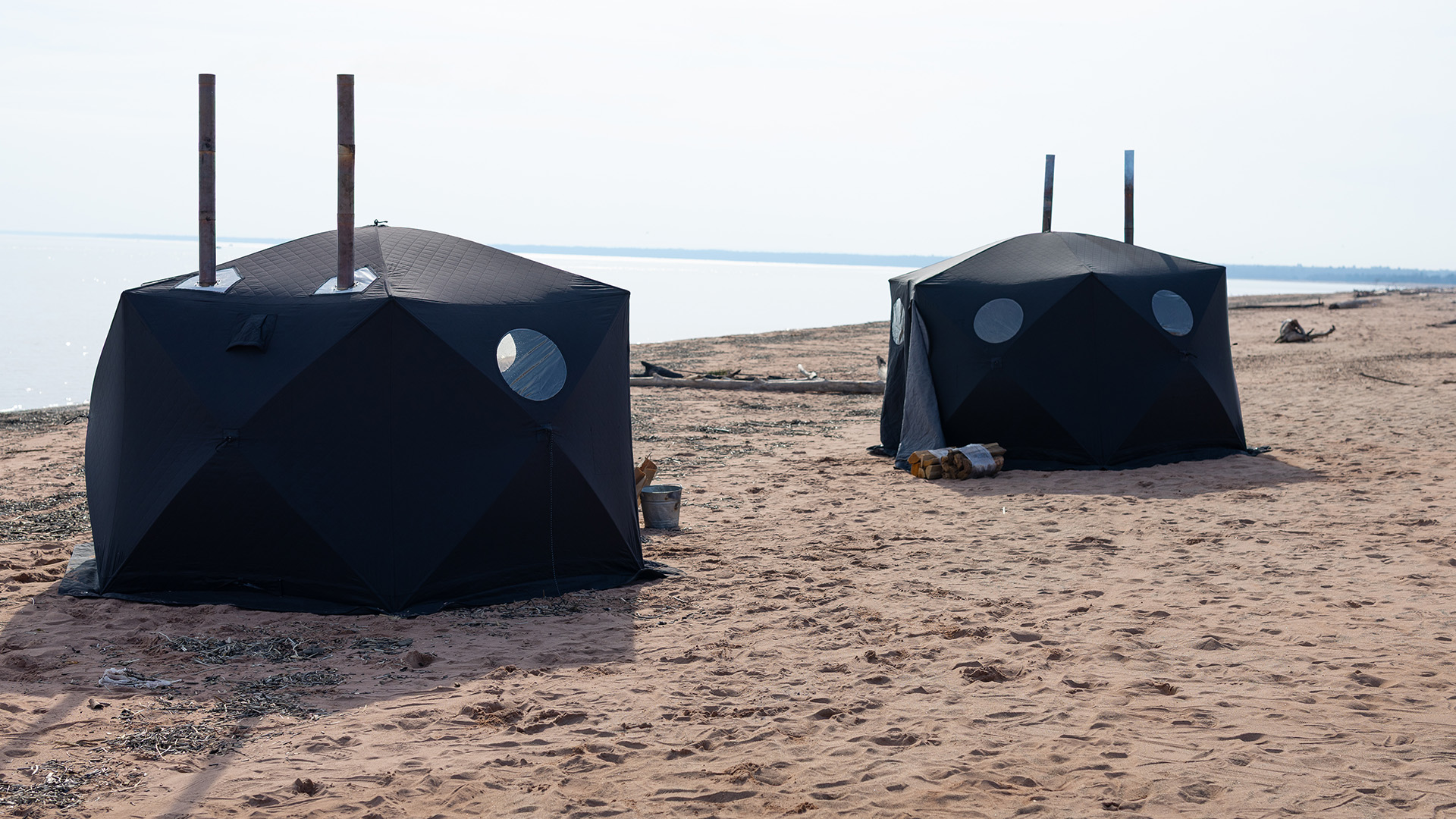 Two black, dome-shaped tents with round windows and pipes on top are set up on a sandy beach near the water’s edge, with some logs and buckets nearby under a clear sky.