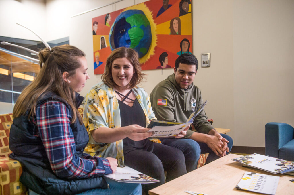 Three young adults sit on a couch, smiling and looking at papers about student resources together in a brightly lit room with colorful artwork on the wall behind them.