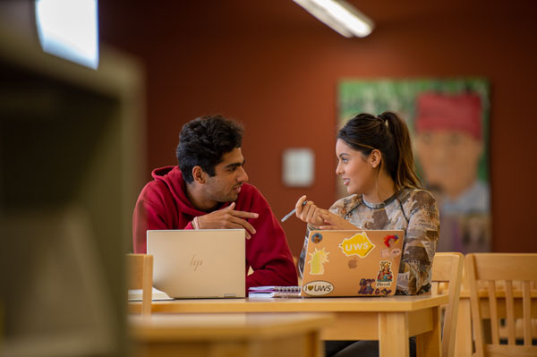Two students sit at a table in a library, talking and smiling, each with a laptop in front of them. The background is blurred, featuring a colorful portrait on the wall—capturing student life at the University of Wisconsin-Superior.