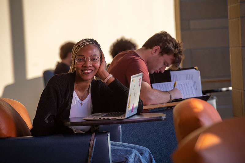 A young University of Wisconsin-Superior student with glasses smiles at the camera while sitting at a desk with a laptop. In the background, a young man is focused on his work, writing in a notebook in a brightly lit study area.