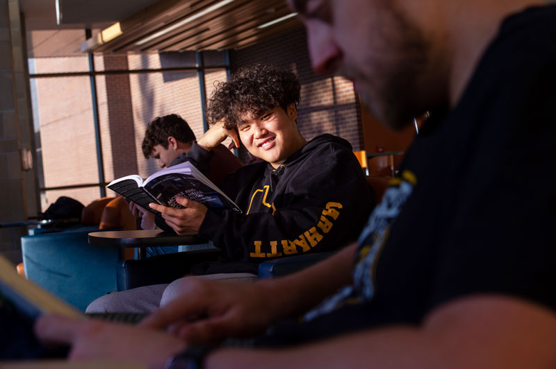 A University of Wisconsin-Superior student with curly hair smiles while holding an open book, sitting in a sunlit room with others nearby, one reading in the foreground and all engaged in their program.