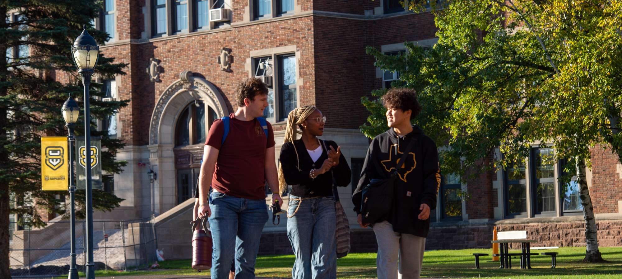 Three students walk together on a sunny campus lawn, chatting and smiling near a large brick academic building, showcasing UW-Superior accessibility with inviting pathways and green trees in the background.