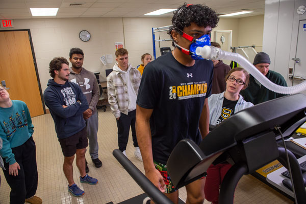 A young man wearing a breathing mask walks on a treadmill for a fitness test while several people from various academic departments, some in athletic wear, watch and take notes in a lab setting.