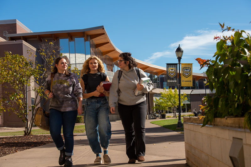 Three students walk together on a sunny University of Wisconsin-Superior campus sidewalk, carrying backpacks and books, with academic buildings, banners, trees, and a lamppost visible in the background.