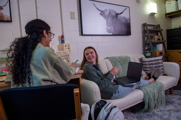 Two students sit and chat in a cozy dorm room during the weekend of welcome; one writes at a desk while the other lounges with a laptop. The room is decorated with a shelf and a framed photo of a cow on the wall.
