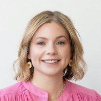 A woman with shoulder-length blonde hair, wearing a pink top and daisy-shaped earrings, smiles in front of a plain white background.
