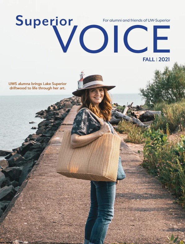 A woman in a straw hat and woven bag stands smiling on a rocky pier by a lake, with a lighthouse behind her. The magazine cover celebrates Alumni achievements and reads Superior VOICE Fall 2021.